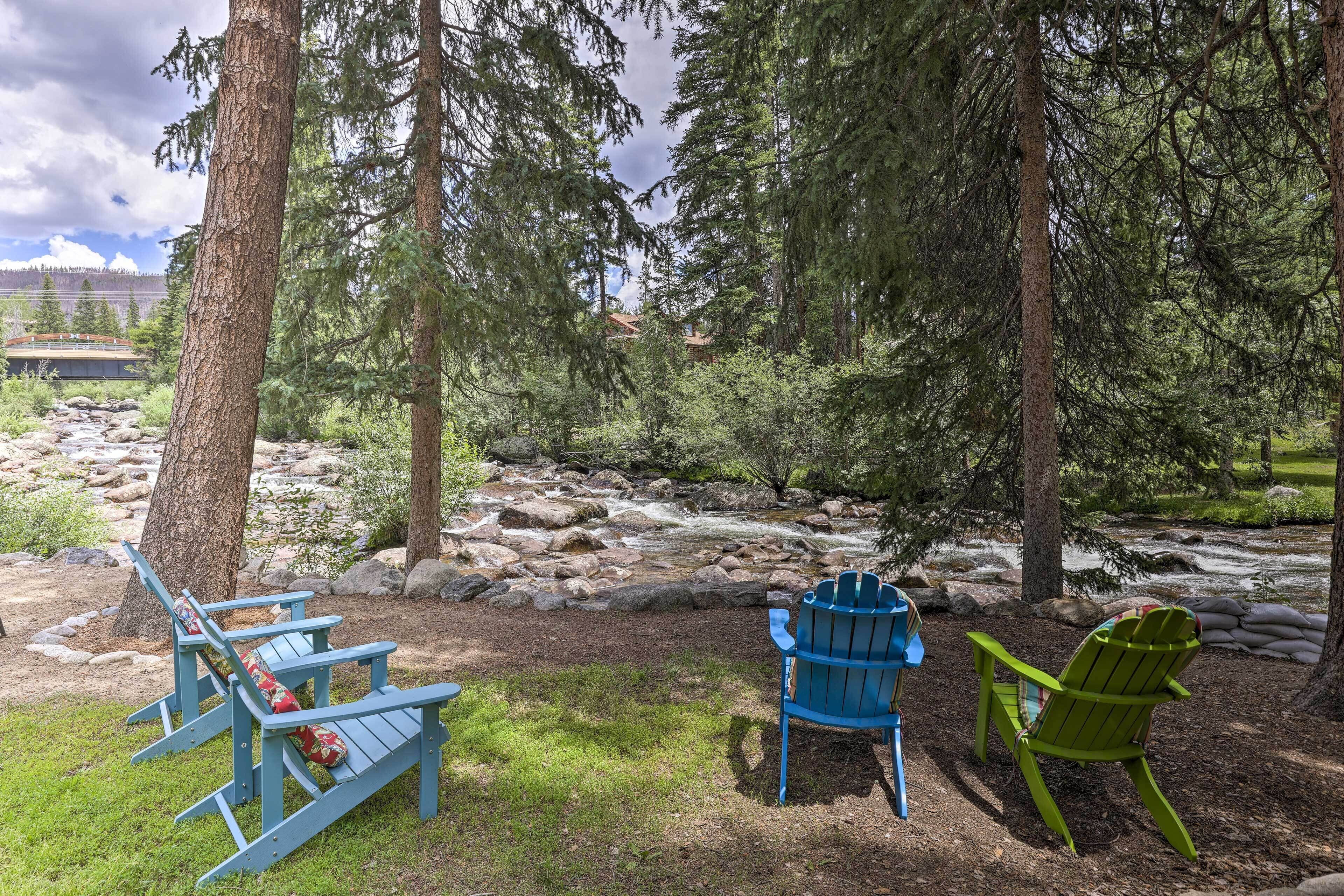 A riverside seating area with Adirondack chairs and a bench, surrounded by trees and a flowing stream.