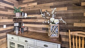 A kitchen with wooden panel walls, a wooden chair, a white cabinet, a coffee maker, a toaster, and a vase with flowers. - Walk to Marina: Rustic Grand Lake Retreat! (Grand Lake)