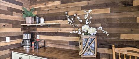 A kitchen with wooden panel walls, a wooden chair, a white cabinet, a coffee maker, a toaster, and a vase with flowers.