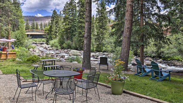 An outdoor seating area with tables and chairs, surrounded by trees and a view of a river and mountains. - Walk to Marina: Rustic Grand Lake Retreat! (Grand Lake)