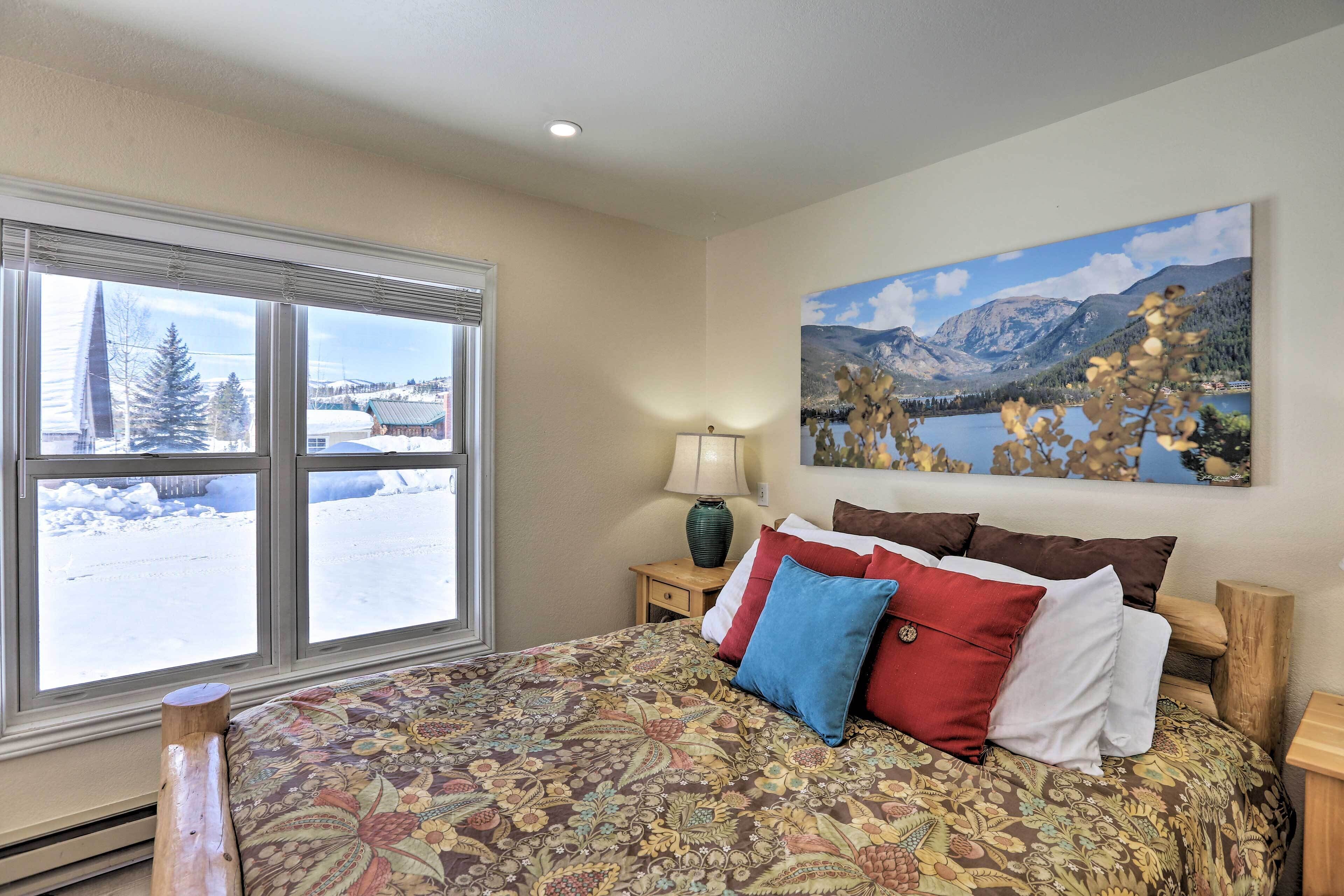 A bedroom with a large window, a bed with patterned bedding, a nightstand with a lamp, and a painting of a mountainous landscape.