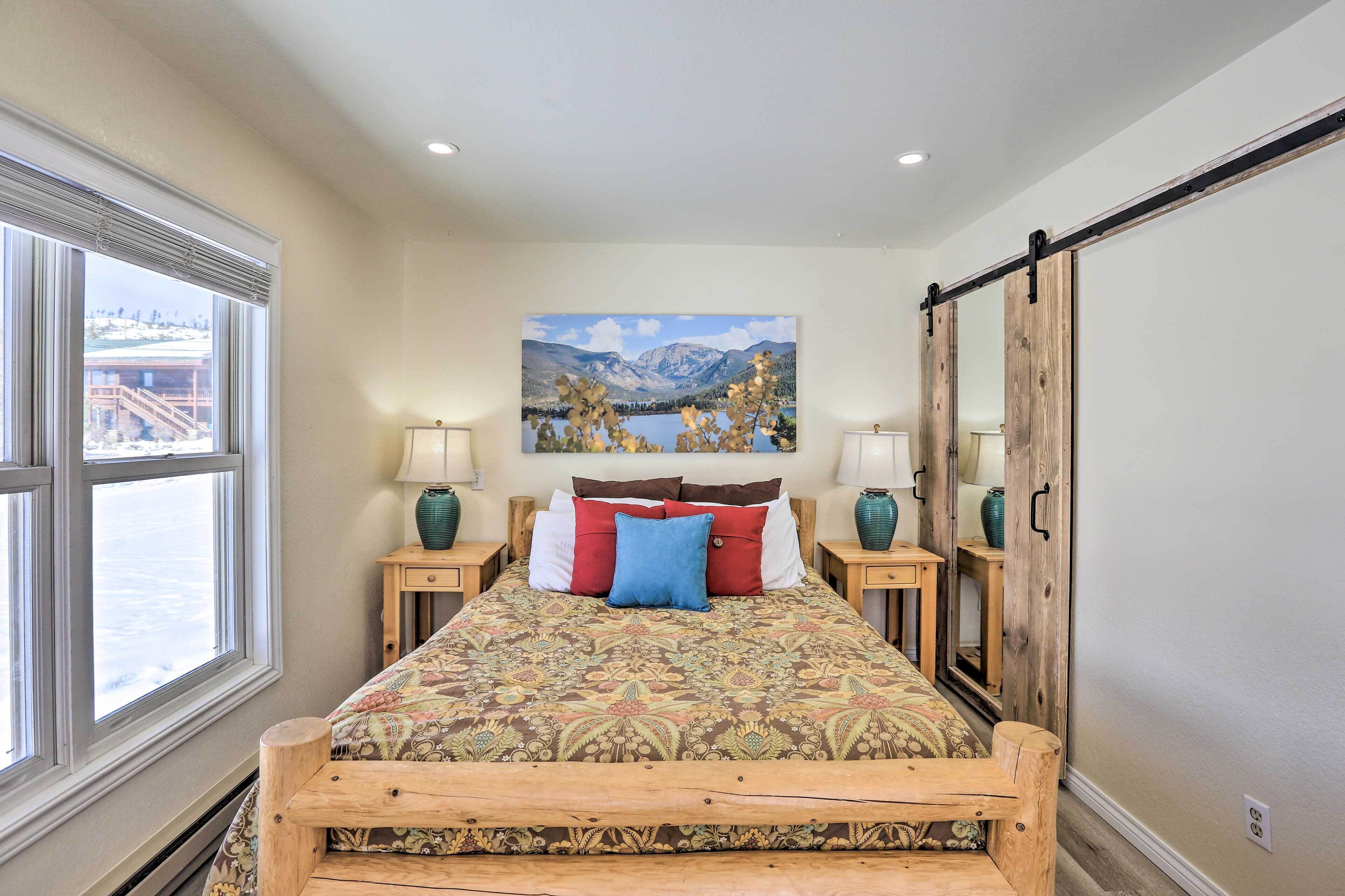 A bedroom with a large bed, a wooden headboard, two bedside tables with lamps, a large window, and a mountain landscape painting on the wall.
