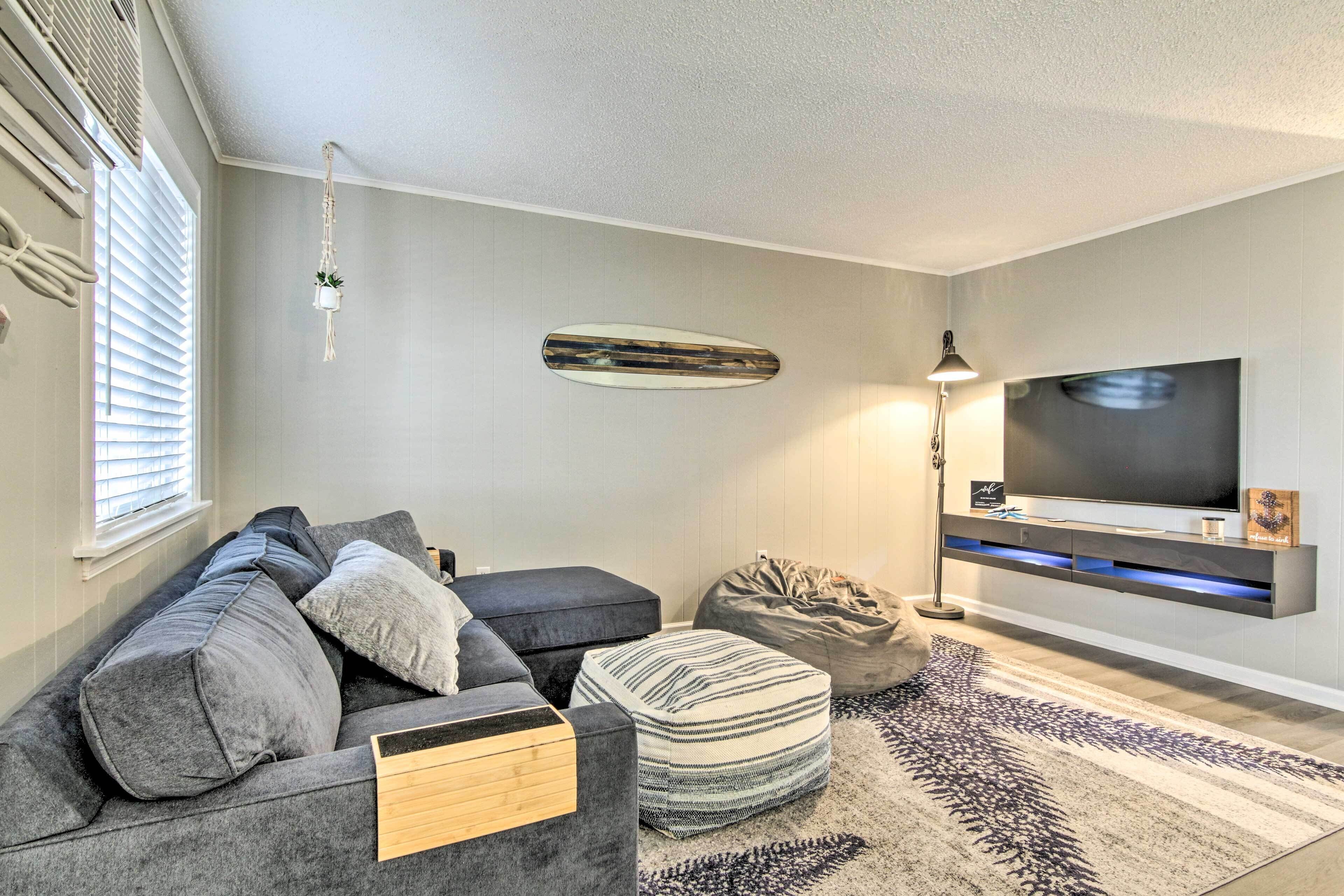 Ein modernes Wohnzimmer mit grauem Sofa, ottoman mit Streifen, Sitzsack, Surfbrett an der Wand und flachbildfernseher auf einer Wandablage.