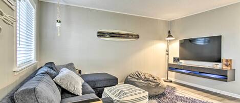 A modern living room with a grey sofa, a striped ottoman, a bean bag, a mounted surfboard, and a flat-screen TV on a wall shelf.