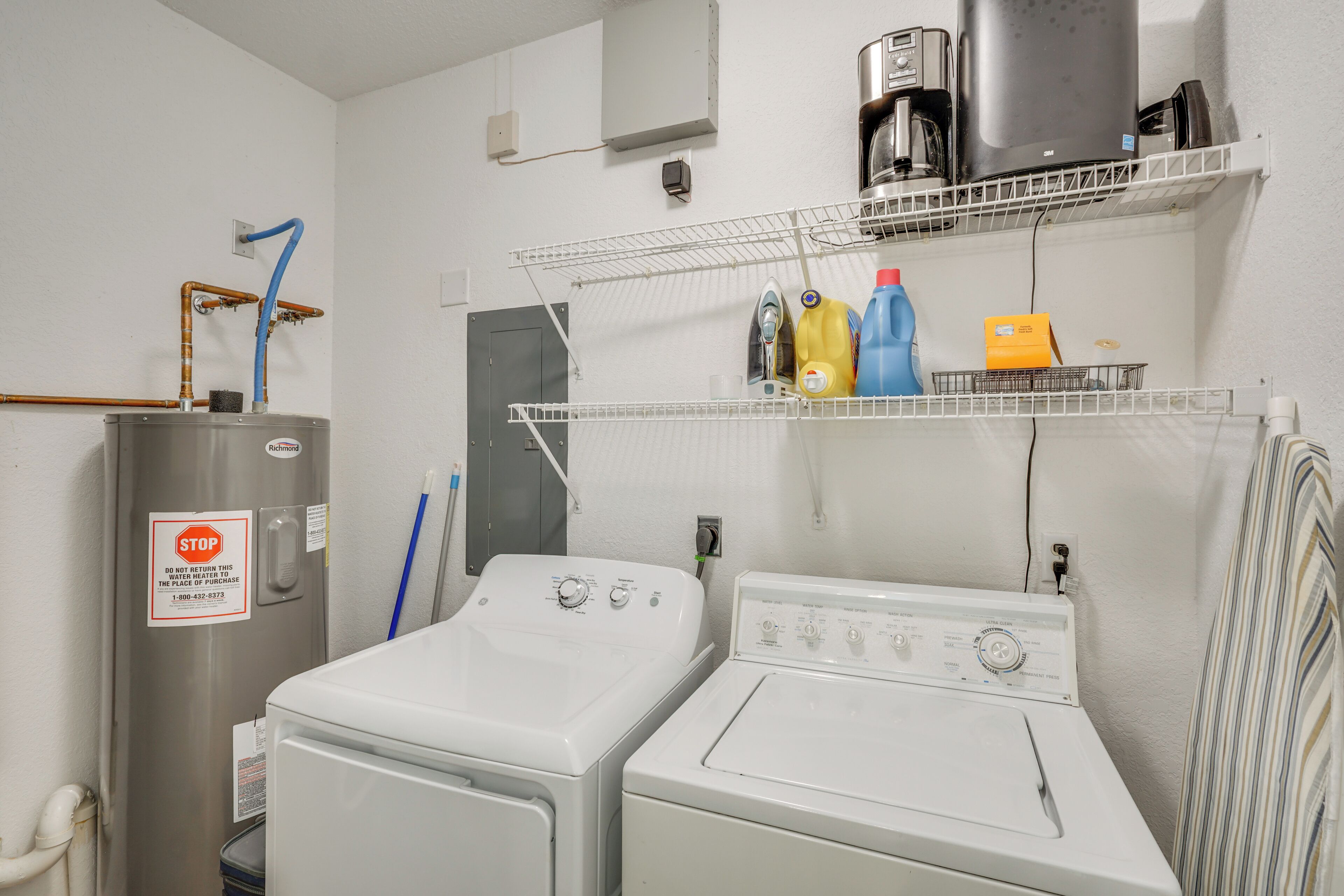 Un espace buanderie équipé de deux machines à laver et deux sèche-linge blancs, d’une machine à café et d’étagères contenant des produits de nettoyage.