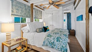 Une chambre avec un plafond en bois, un lit recouvert d’une literie à motifs floraux, une table de chevet avec des livres et une fenêtre avec des rideaux.