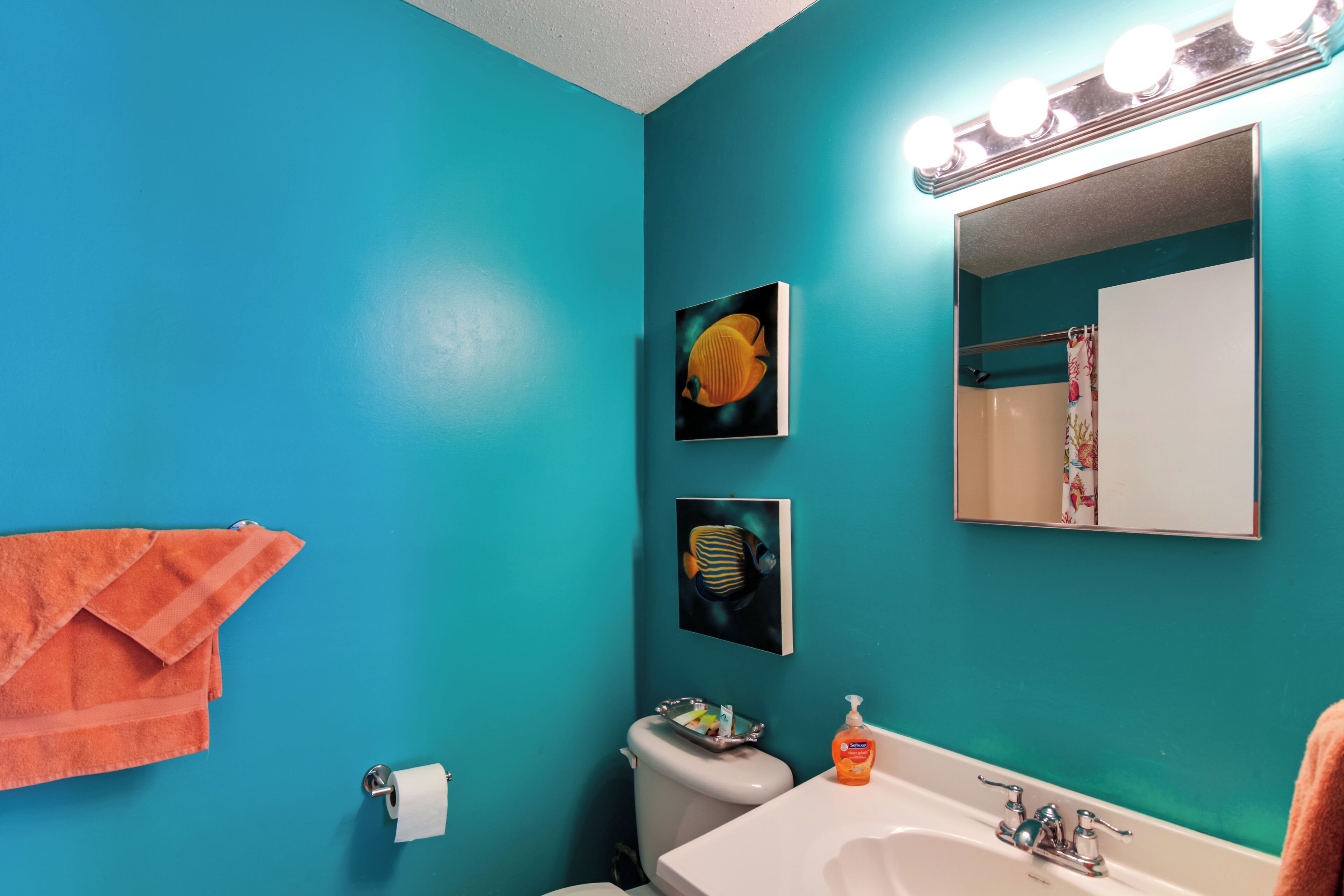 A bathroom with teal walls, a white sink, a toilet, and a mirror with lights above it.