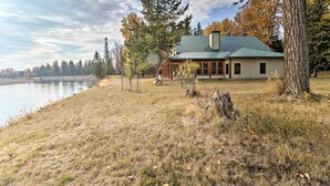 A cabin by a lake with a metal roof, surrounded by trees and a grassy field. - Kalispell Riverfront Home by Glacier National Park (Kalispell)