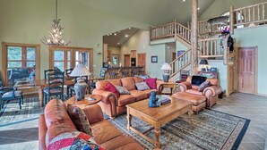 A spacious living room with a large sectional sofa, a wooden coffee table, and a chandelier. A staircase with a wooden railing leads to an upper level. - Kalispell Riverfront Home by Glacier National Park (Kalispell)
