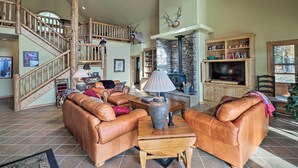 A spacious living room with a high ceiling, a wooden staircase, leather sofas, a fireplace, and a television. - Kalispell Riverfront Home by Glacier National Park (Kalispell)
