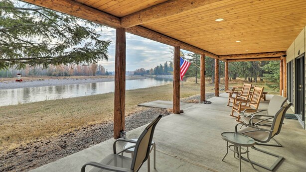 A covered outdoor seating area with chairs and tables, overlooking a body of water and forested area. - Kalispell Riverfront Home by Glacier National Park (Kalispell)