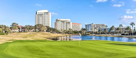 A golf course with a water hazard, surrounded by high-rise buildings and palm trees.