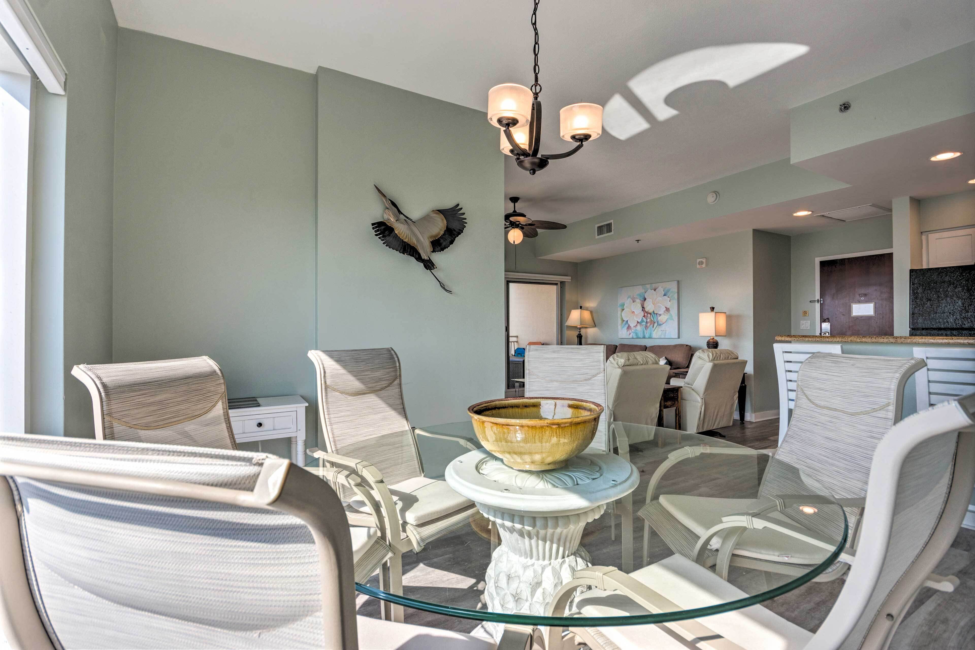 Un coin repas avec une table en verre, des chaises blanches, un lustre et une décoration murale représentant des oiseaux.