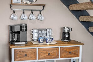 Eine Küchenzeile mit einer Kaffeemaschine, einem Wasserkocher und einer Kaffeekanne auf der Arbeitsfläche. Darüber hängen Tassen an einem Ablagegestell.