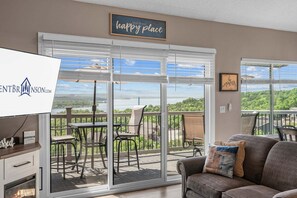 A room with a sliding glass door leading to a balcony with a view of a lake and trees. There is a couch with a cushion and a television on a stand.