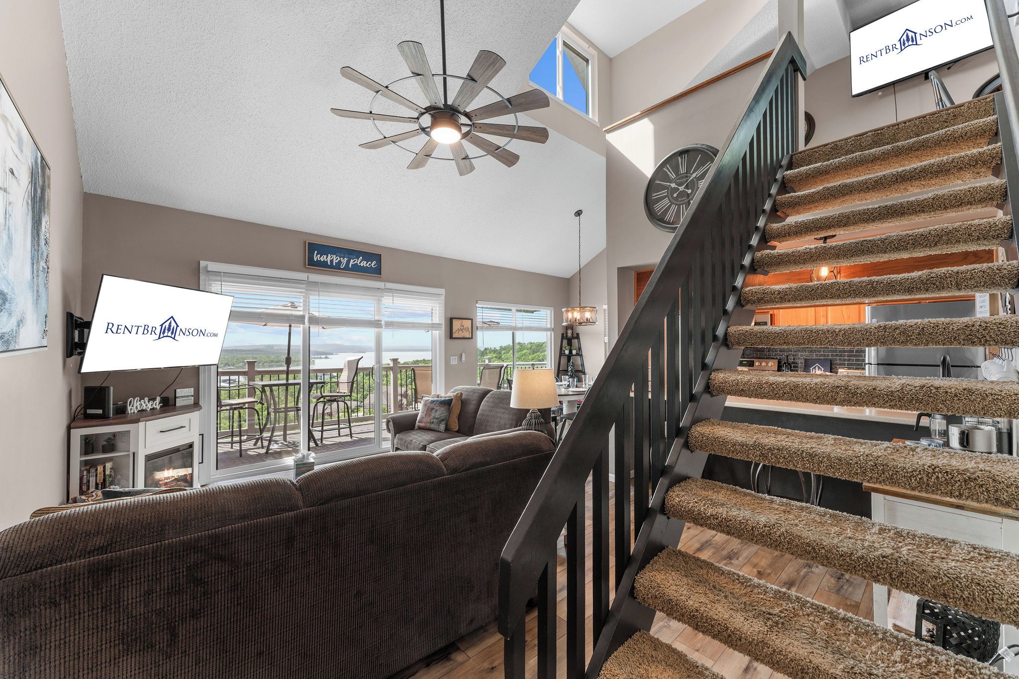 A spacious living room with a large window, a ceiling fan, a staircase with a unique carpet design, and a dining area.