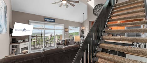 A spacious living room with a large window, a ceiling fan, a staircase with a unique carpet design, and a dining area.