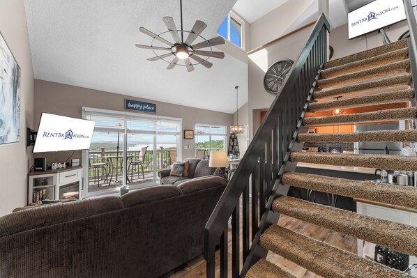 A spacious living room with a large window, a ceiling fan, a staircase with a unique carpet design, and a dining area.