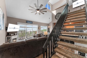 A spacious living area with a large window, a ceiling fan, a staircase with a unique carpeted design, and a sign indicating a rental company.