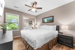 A bedroom with a bed, bedside tables, a nightstand, a ceiling fan, a window with blinds, and a painting on the wall.