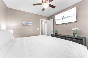 Ein Schlafzimmer mit einem Bett, einem Deckenventilator, einem an der Wand montierten Fernseher und einer Tür zu einem anderen Raum.