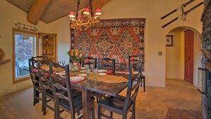 Un comedor con una mesa de madera dispuesta para seis personas, rodeada de sillas. Un candelabro cuelga del techo y un tapiz con motivos decorativos adorna la pared detrás de la mesa.