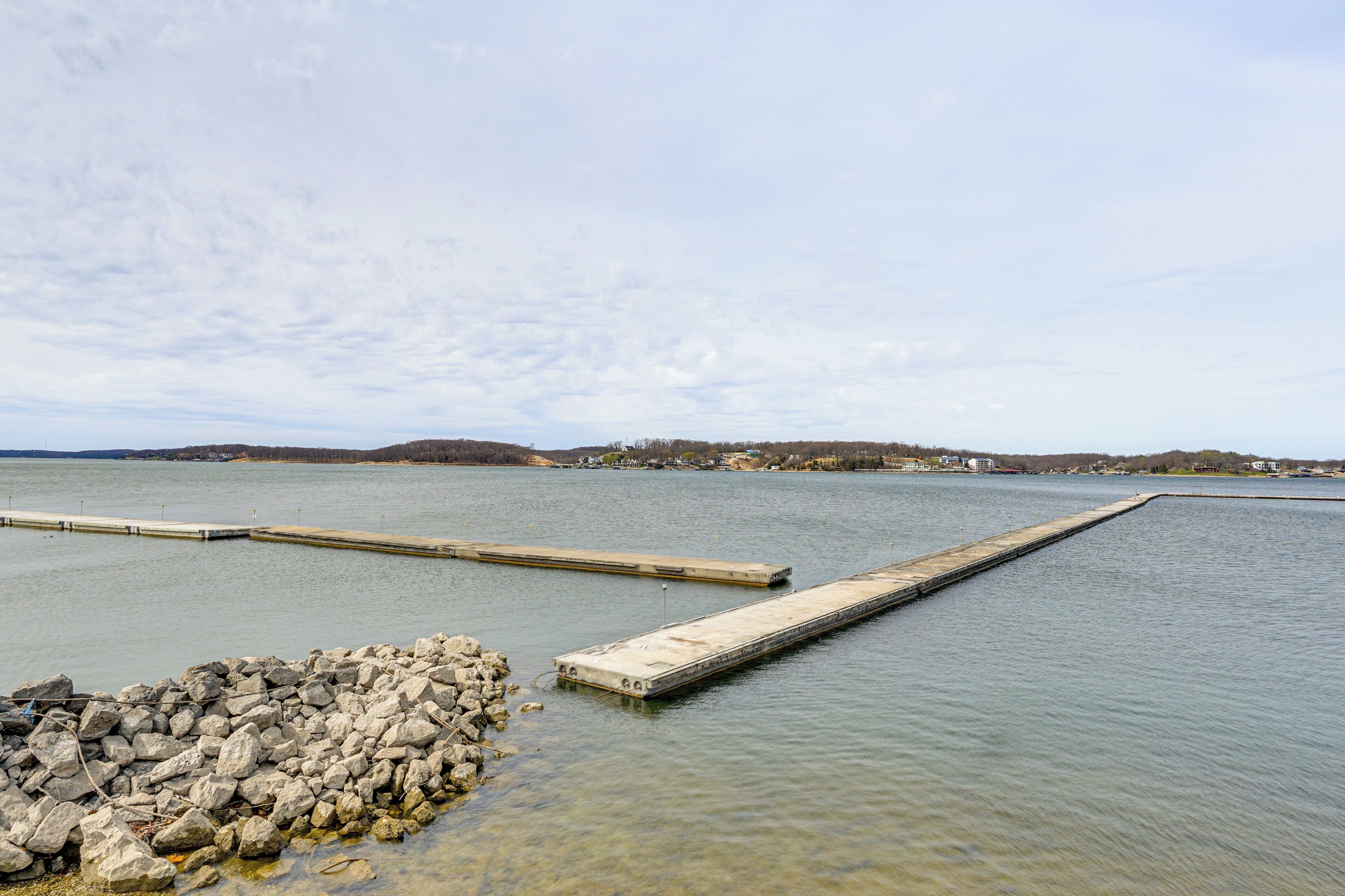 Un lago tranquilo con un muelle que se adentra en el agua, rodeado por una orilla rocosa y colinas al lejano.