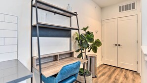 A compact room with a wooden desk, a blue chair, a metal shelving unit, a potted plant, a trash bin, and a white wardrobe.