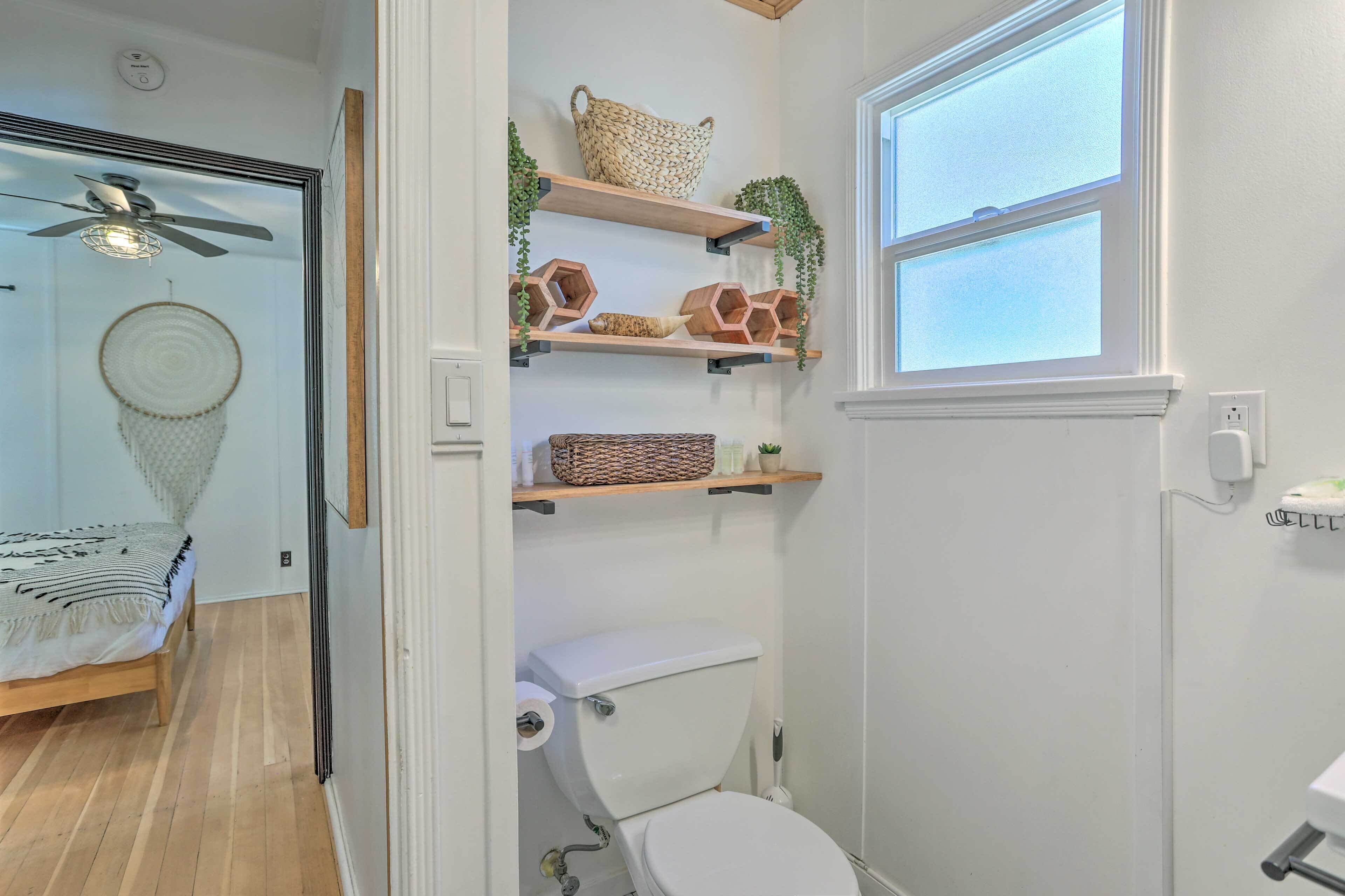 A bathroom with a white toilet, wooden shelves with decorative items, a ceiling fan, and a window.