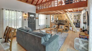 Uma sala de estar ampla com teto alto, vigas de madeira, um sofá, poltronas, lareira e escada que leva a um andar superior.