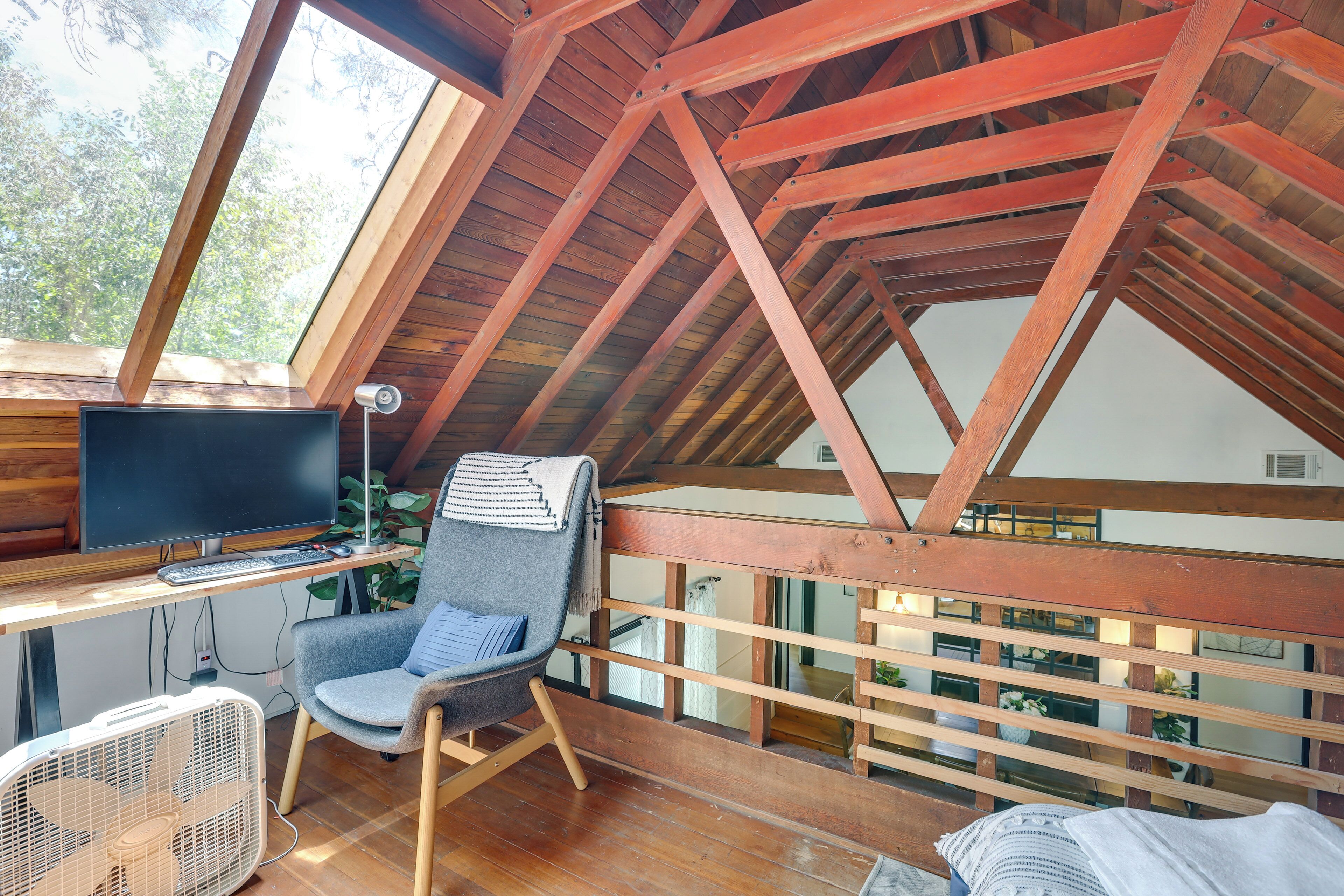 Une pièce au plafond haut, avec une structure en bois, un bureau équipé d’un ordinateur, une chaise et un ventilateur.