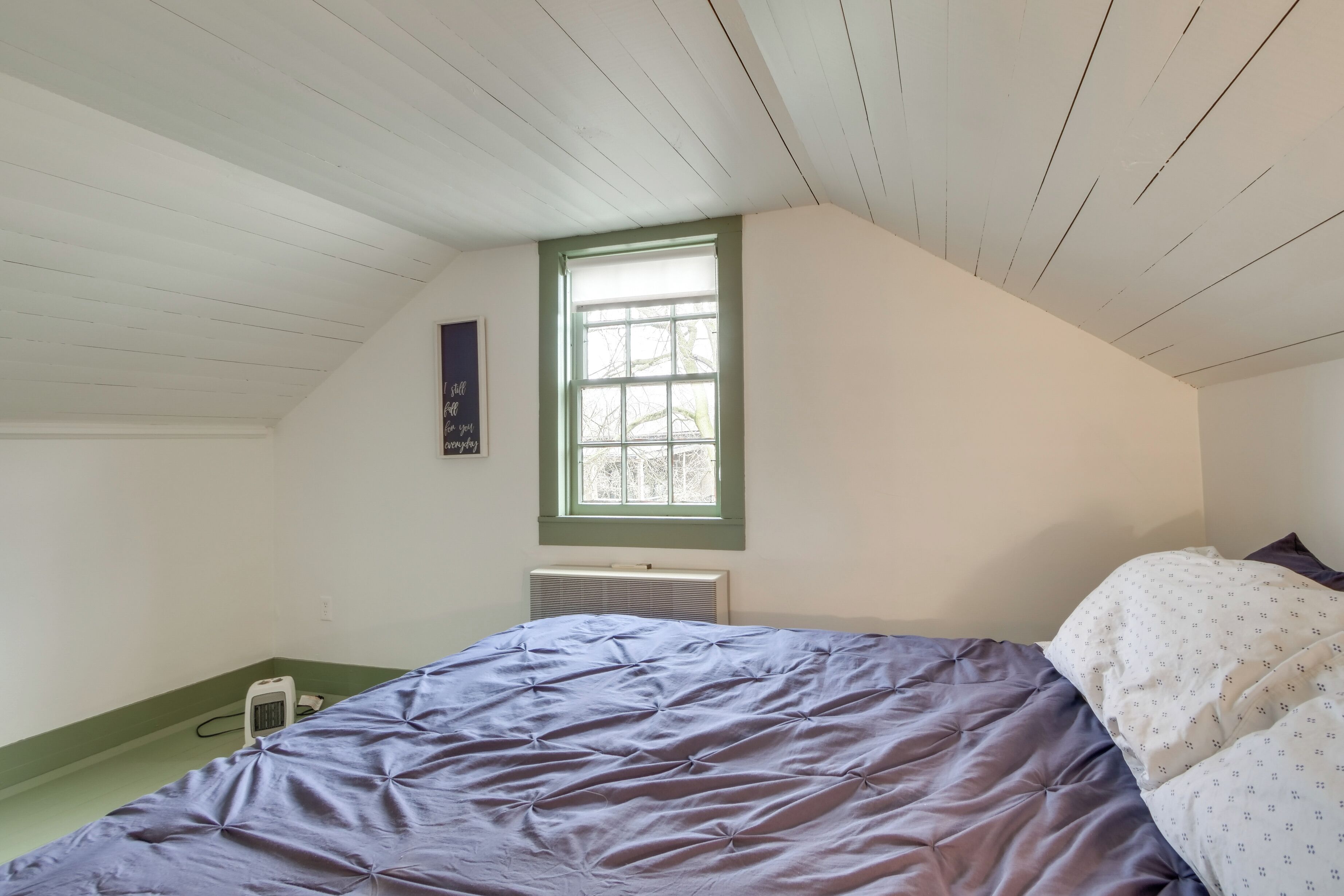 A bedroom with a sloped ceiling, a window, a bed with a blue bedspread, and a white radiator.