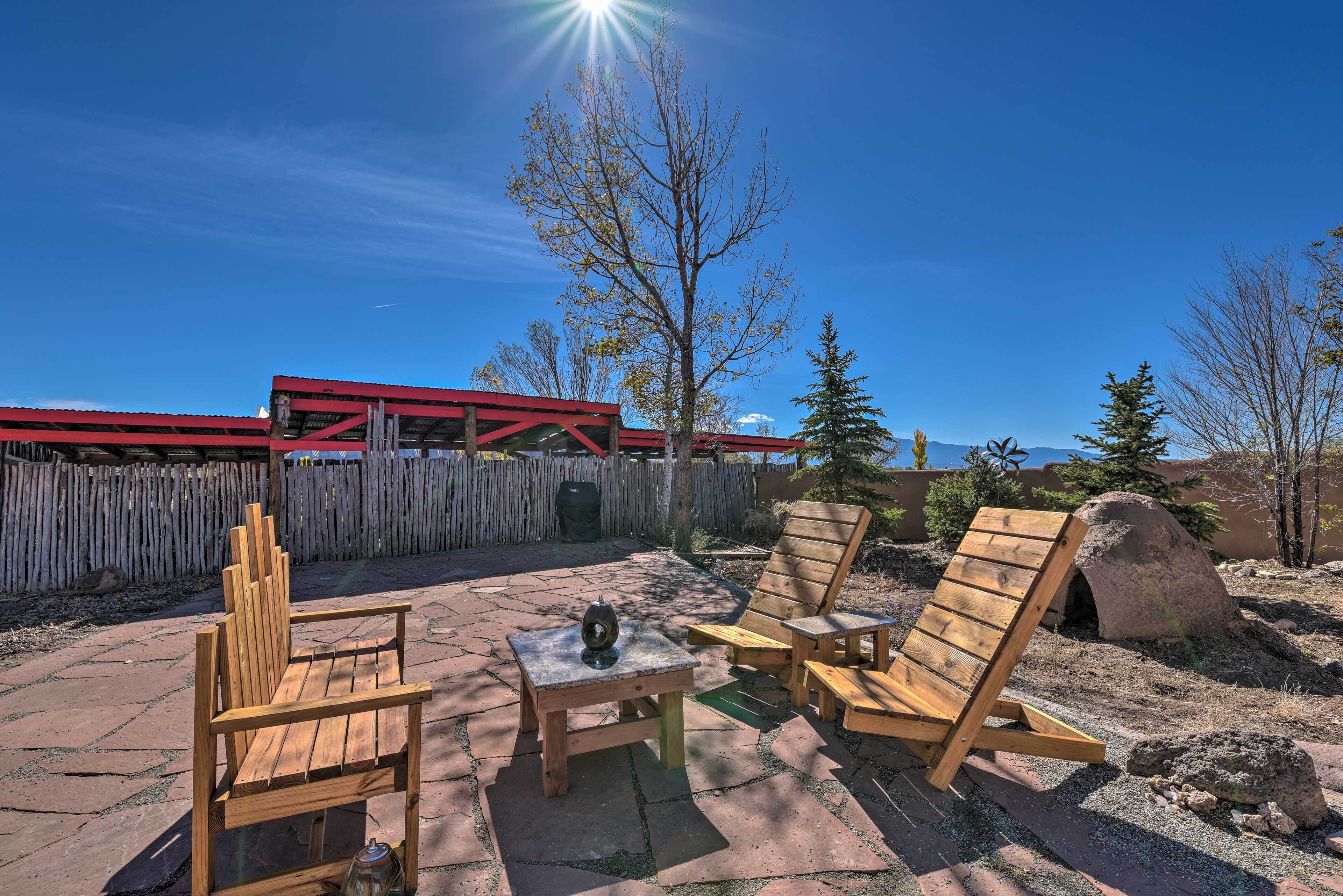 Une terrasse extérieure meublée de mobilier en bois, comprenant des chaises et une table, avec en arrière-plan un bâtiment rouge et un ciel bleu dégagé.
