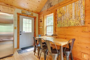 A cabin interior with a wooden dining table, four chairs, a refrigerator, and a painting of a forest. - Rustic Cabin w/ Hot Tub - 7 Miles to Hocking Hills (Logan)