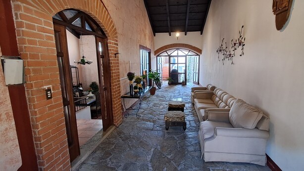 A spacious corridor with a stone floor, a brick wall, and a white wall with decorative elements. There are potted plants and a beige sofa along the corridor. - The Orchid Hotel (Antigua Guatemala)
