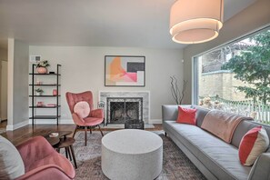 Een moderne woonkamer met een open haard, een ronde salontafel, een bank met kussens, een roze fauteuil en een boekenkast.