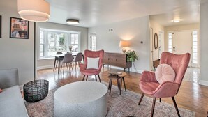 A modern living room with a round coffee table, two pink armchairs, a dining area with a table and chairs, and a large window providing natural light.