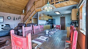 A spacious dining area with a long wooden table, red chairs, and a chandelier. A kitchen is visible in the background with wooden cabinets and a stove. - Ruidoso Home w/ Hot Tub, Mtn Views & Game Room! (Ruidoso)