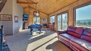 A spacious room with a pool table, leather sofa, and large windows offering a view of the outdoors. - Ruidoso Home w/ Hot Tub, Mtn Views & Game Room! (Ruidoso)