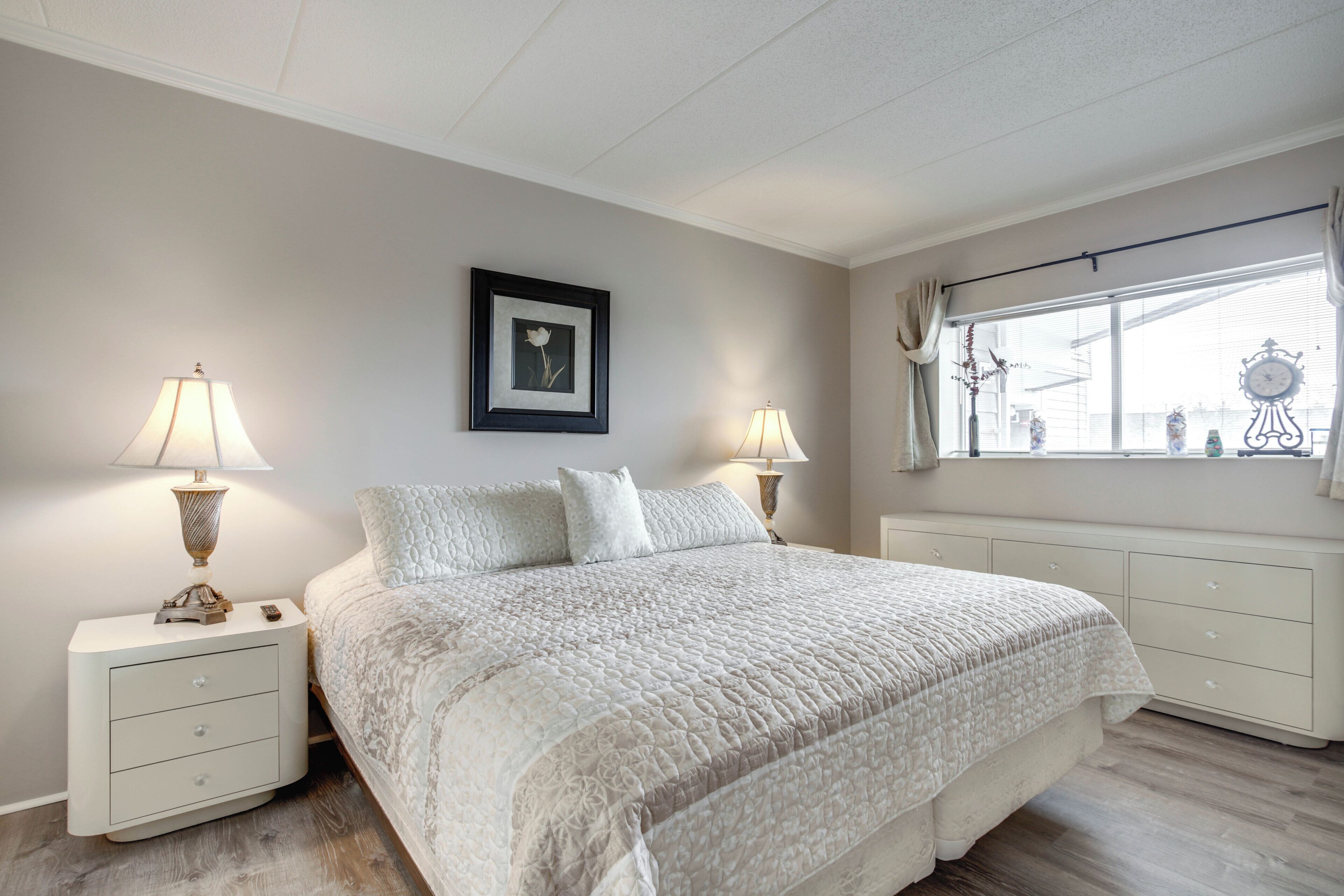 Un dormitorio con una cama grande, dos mesitas de noche con lámparas, un cuadro enmarcado en la pared y una ventana con cortinas.