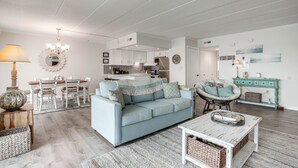 Een ruime woonkamer met een lichtblauwe bank, een eethoek met witte stoelen, en een keuken op de achtergrond.