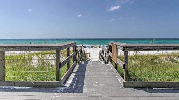 Une promenade en bois mène à une plage où l’eau est d’un bleu cristallin et où quelques parasols sont installés.