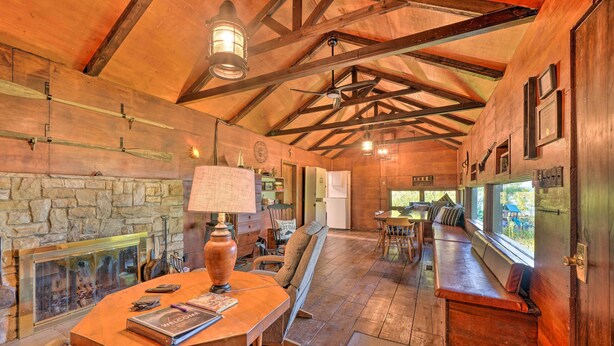 A rustic interior with a stone fireplace, wooden beams, and a large window overlooking a scenic view. - Custom-built Lakefront Cottage w/ Spacious Yard! (Westford)