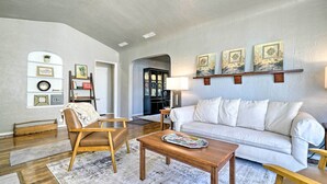 A living room with a white sofa, wooden coffee table, and a chair. There are wall-mounted shelves with decorative items and a door leading to another room. - El Paso Home w/ Backyard + Outdoor Fireplace! (El Paso)