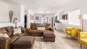 Amplia sala de estar con sofá modular, un sillón amarillo y una mesa de centro. Se ve un área de cocina al fondo, con un refrigerador y armarios.