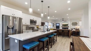 Una cocina moderna con gabinetes blancos, electrodomésticos de acero inoxidable, una isla central con taburetes altos e iluminación colgante.