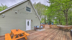 Una terraza de madera con mesas de picnic y mobiliario exterior, rodeada por una valla de madera y árboles.