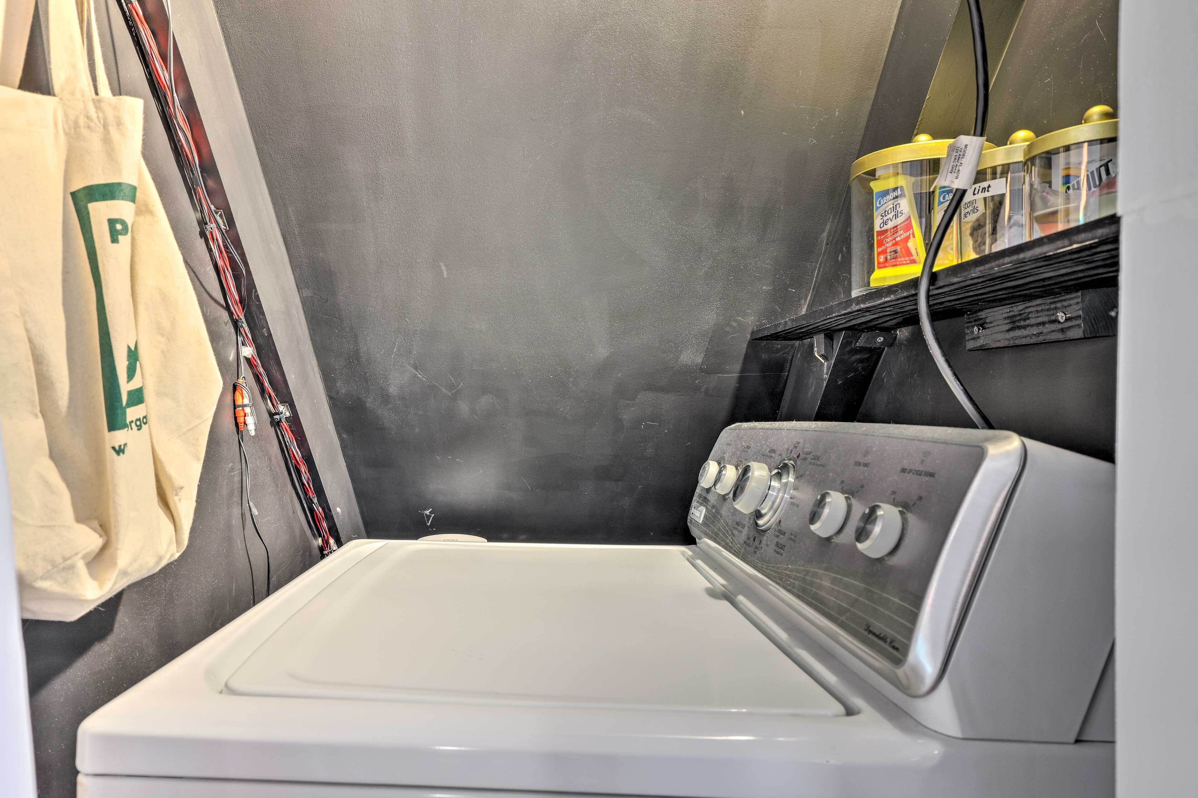 A white washing machine with control knobs, a beige tote bag, and detergent bottles on a shelf.