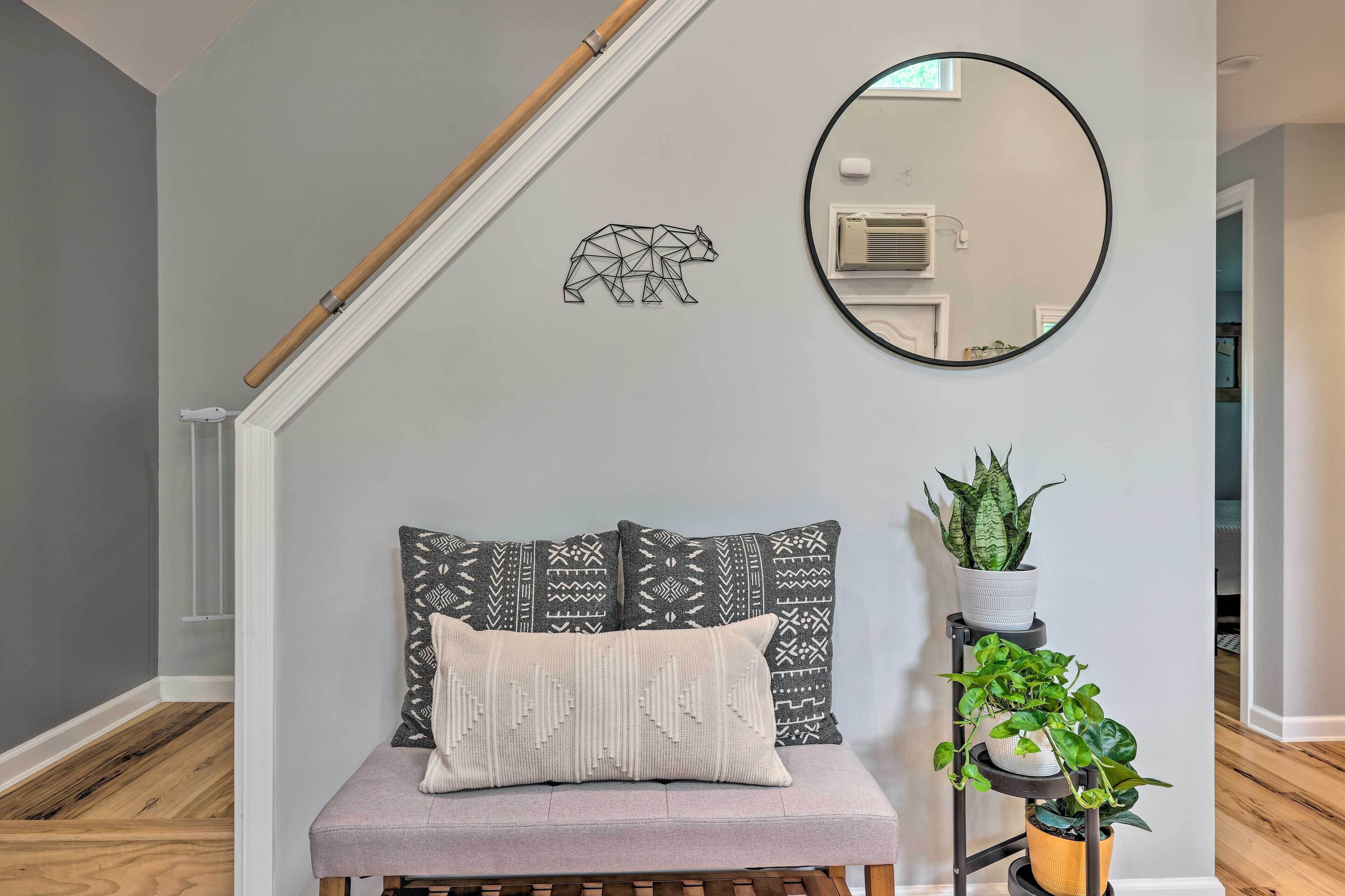 A room with a grey wall, a wooden bench with patterned cushions, a mirror, and a plant stand with green plants.