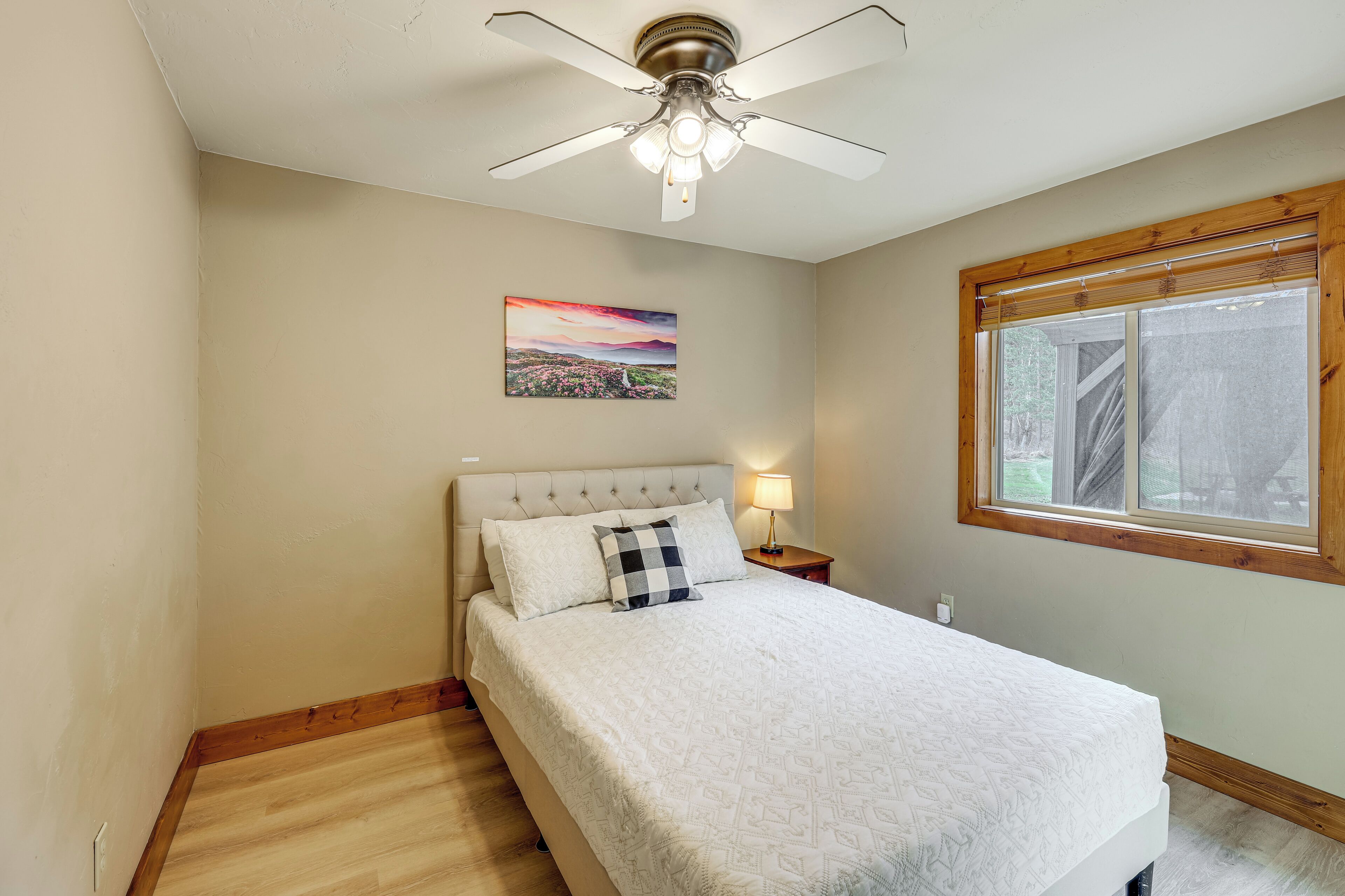 A bedroom with a bed, a ceiling fan, a nightstand, a lamp, a window, and a wall-mounted painting.